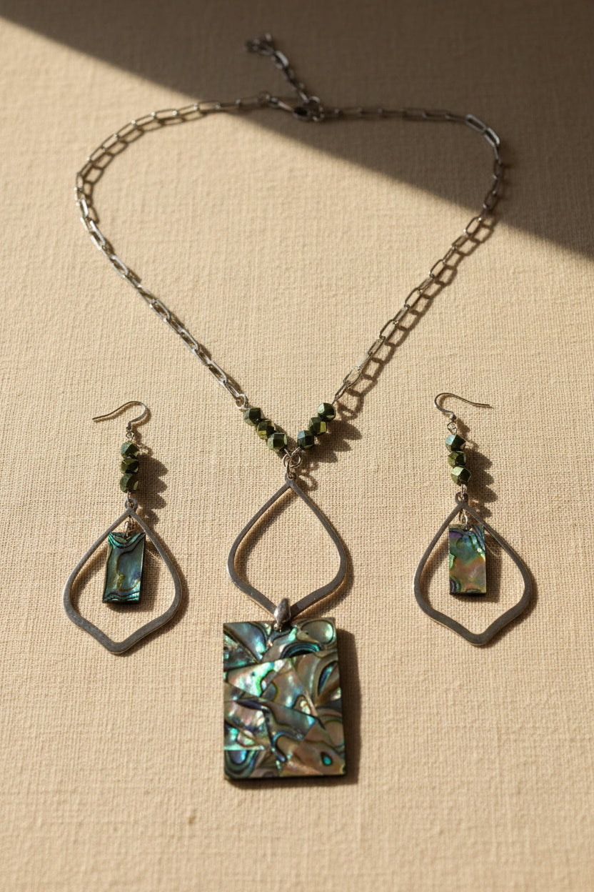 A jewelry set featuring a silver-tone chain necklace and matching drop earrings with iridescent abalone shell inlays and dark green faceted beads, displayed on a light surface.